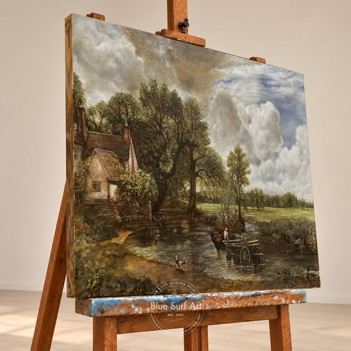 A landscape painting on canvas displayed on a wooden easel in a gallery setting. The painting depicts a rustic cottage beside a river with trees, a boat, and dramatic clouds.