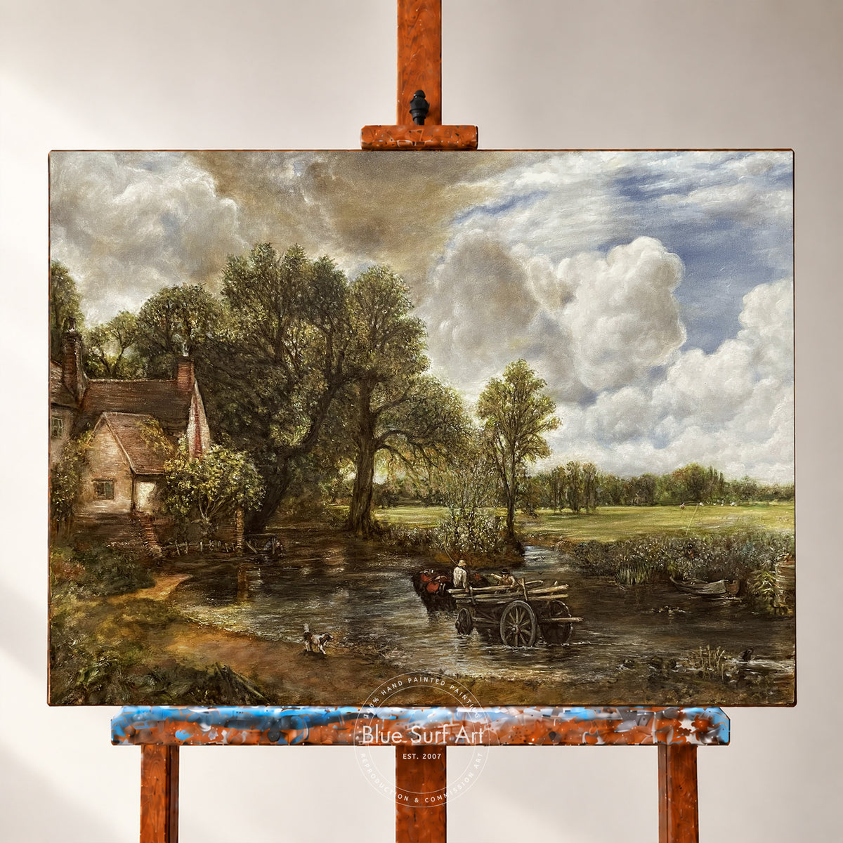 A landscape painting on canvas displayed on an easel. The artwork depicts a rustic countryside scene with a small cottage, large trees, a river or stream, and a cloudy sky.